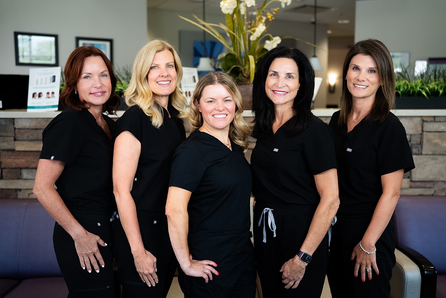 Five women in black scrubs smiling together.